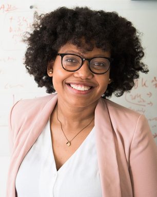 a woman with glasses smiling in front of a whiteboard.